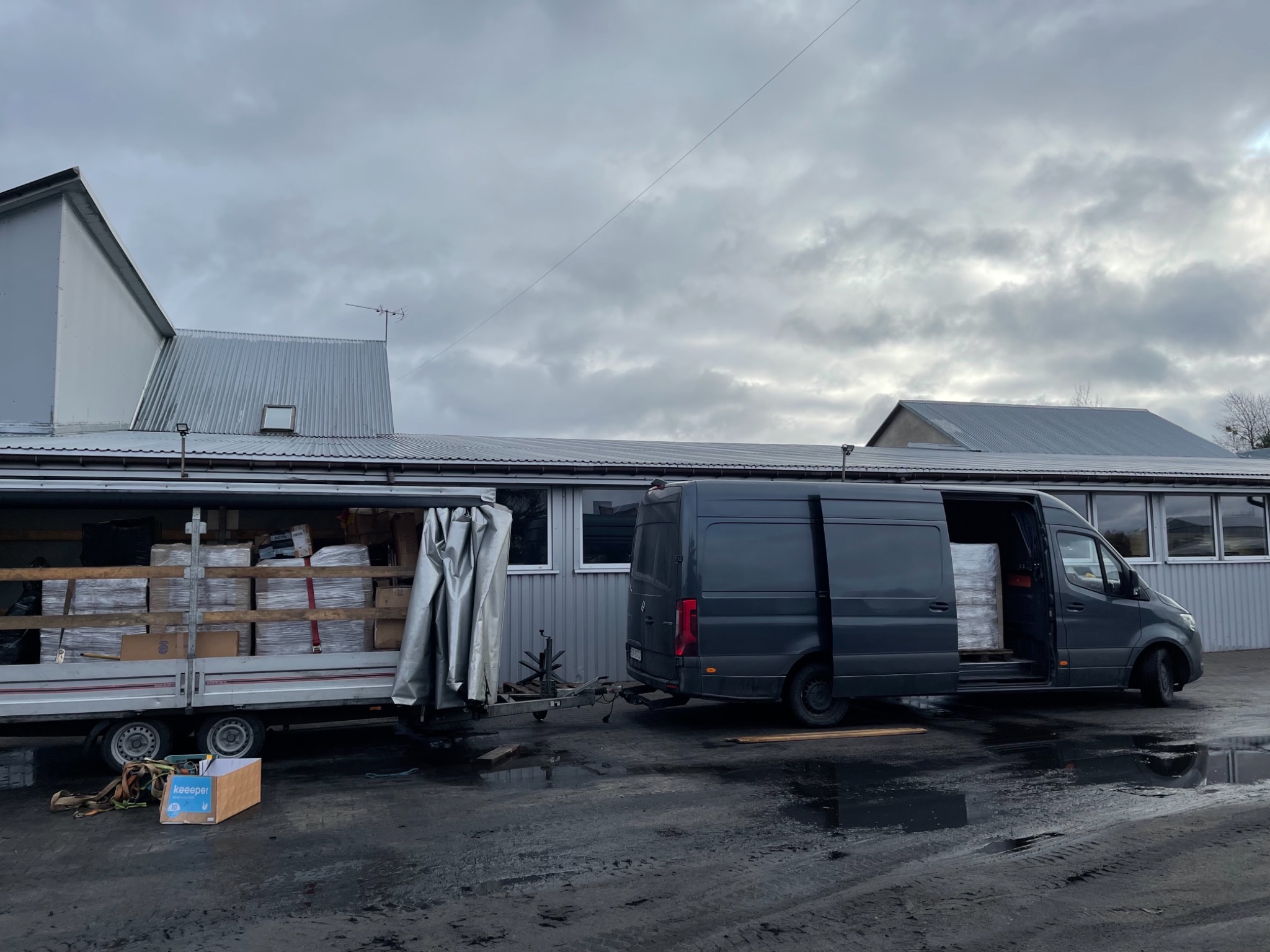 Szary bus z otwartymi drzwiami bocznymi, częściowo załadowany paczkami, zaparkowany obok przyczepy z plandeką, również załadowanej towarem. Mokry asfalt i budynek w tle, pochmurne niebo.