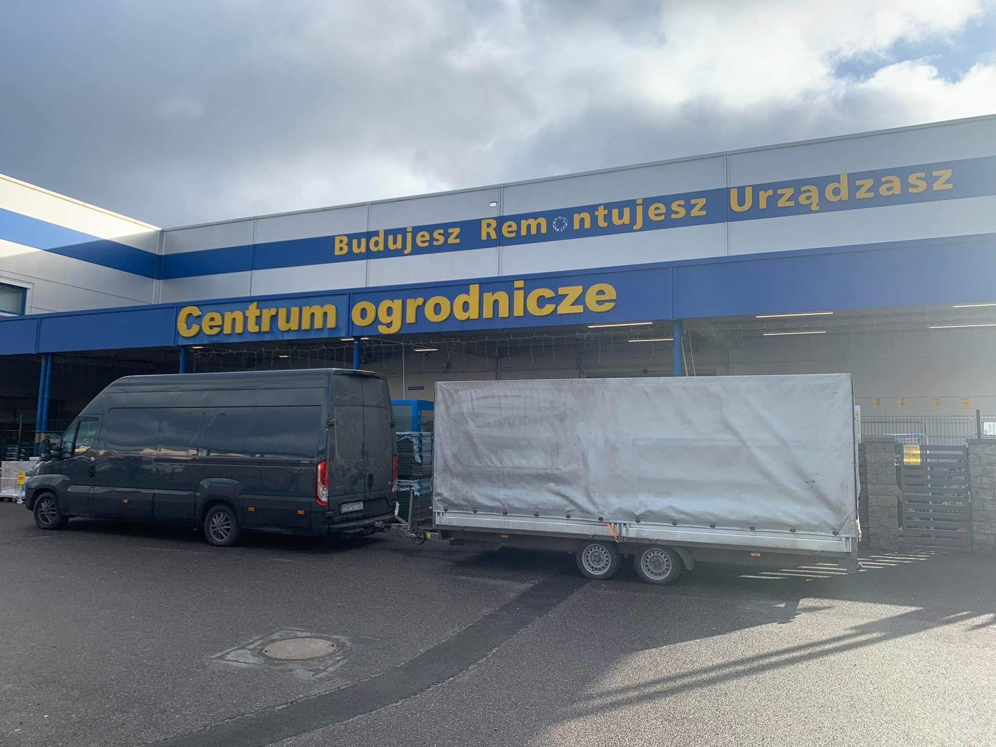Czarny bus z przyczepą pod zadaszeniem sklepu ogrodniczego z napisem: Budujesz, Remontujesz, Urządzasz.