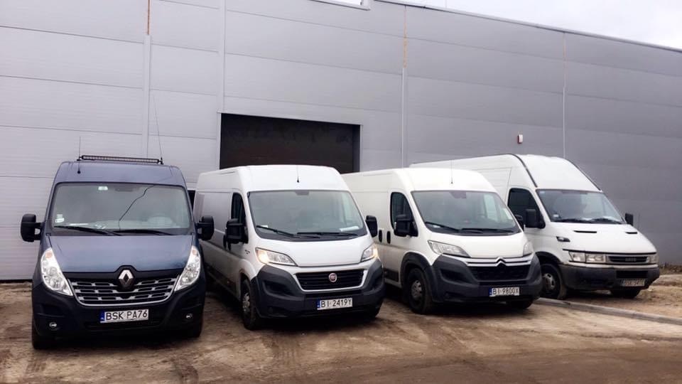 Flota czterech busów transportowych różnych marek (Renault, Fiat, Citroen, Iveco) zaparkowana przed szarym budynkiem magazynowym.