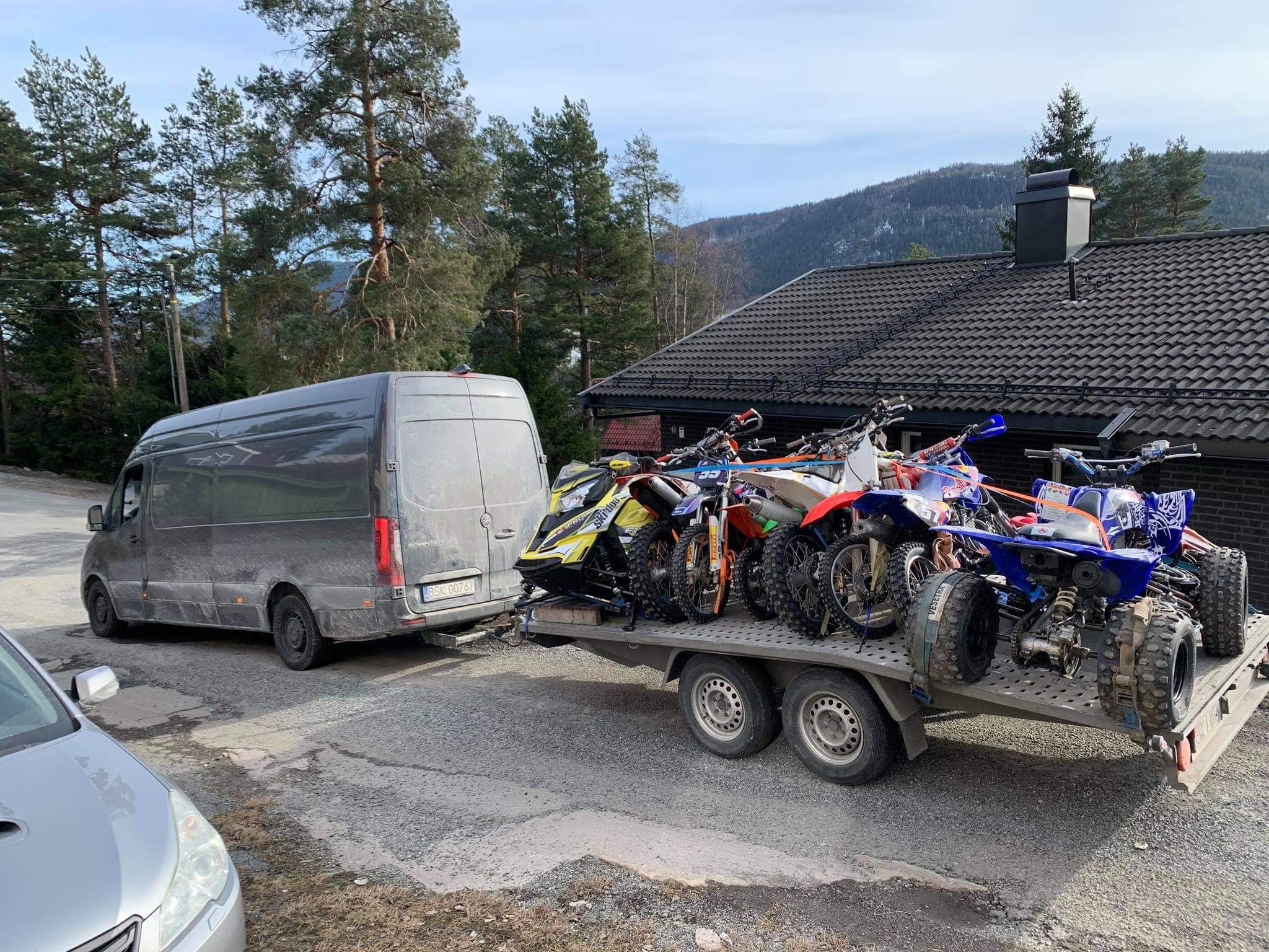Szary, lekko zabrudzony bus ciągnie przyczepę z kilkoma motocyklami i quadami na tle domu i górskiego krajobrazu.
