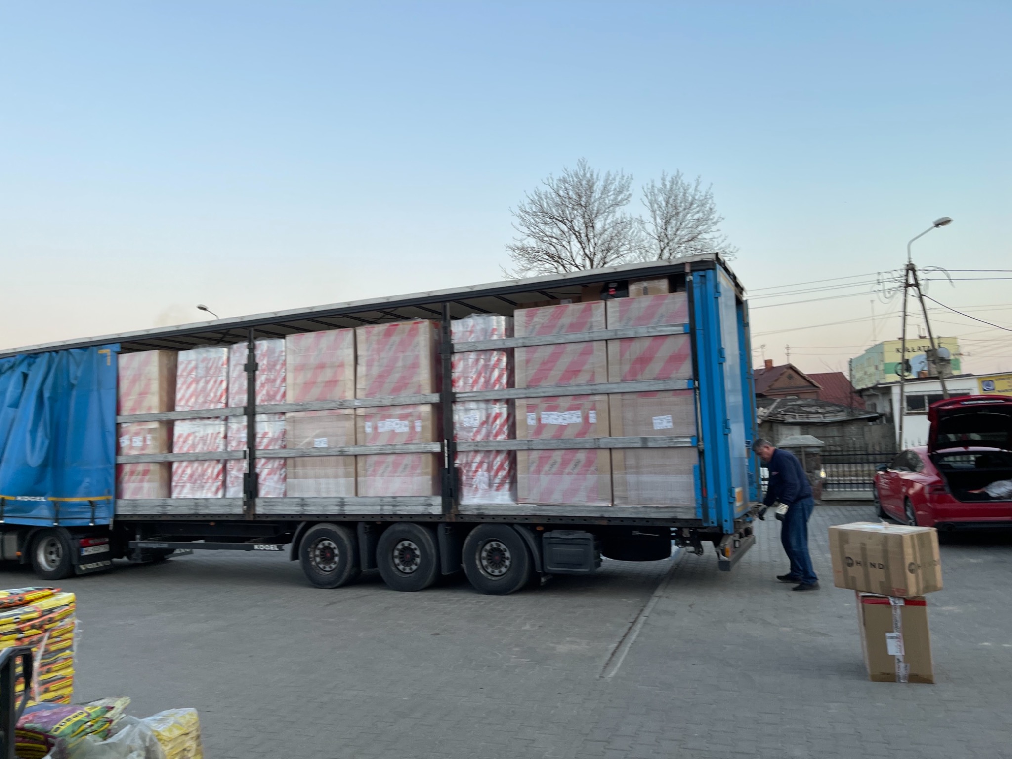 Ciężarówka z naczepą wypełniona po brzegi zapakowanymi kartonami, rozładunek w Sokółce, w tle czerwony samochód osobowy z otwartym bagażnikiem i sylwetka pracownika w trakcie pracy.