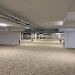 Wnętrze parkingu podziemnego z nawierzchnią z kostki brukowej w odcieniach beżu, filary oznaczone czarno-żółtymi pasami, widoczne oznaczenia miejsc parkingowych i sufit z instalacjami.