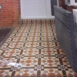 Ułożona, wzorzysta posadzka z płytek ceramicznych przed wejściem do domu z białą stolarką drzwiową i fragmentem muru z czerwonej cegły.