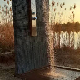 Zewnętrzny prysznic z deszczownicą, obłożony mozaiką, na tle jeziora i trzcin w promieniach zachodzącego słońca.