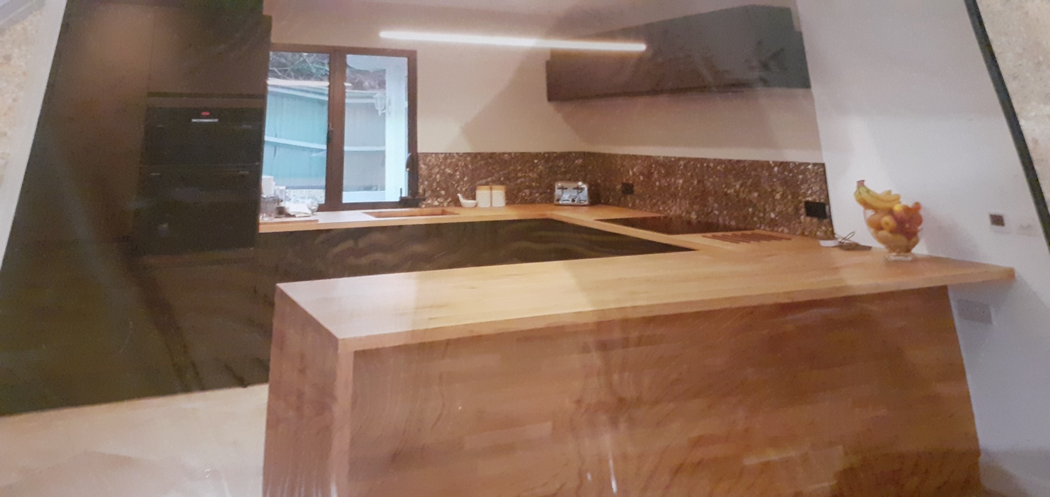 Nowoczesna kuchnia z drewnianym blatem, mozaikową ścianą nad blatem i owocami w szklanej misie na końcu wyspy kuchennej.