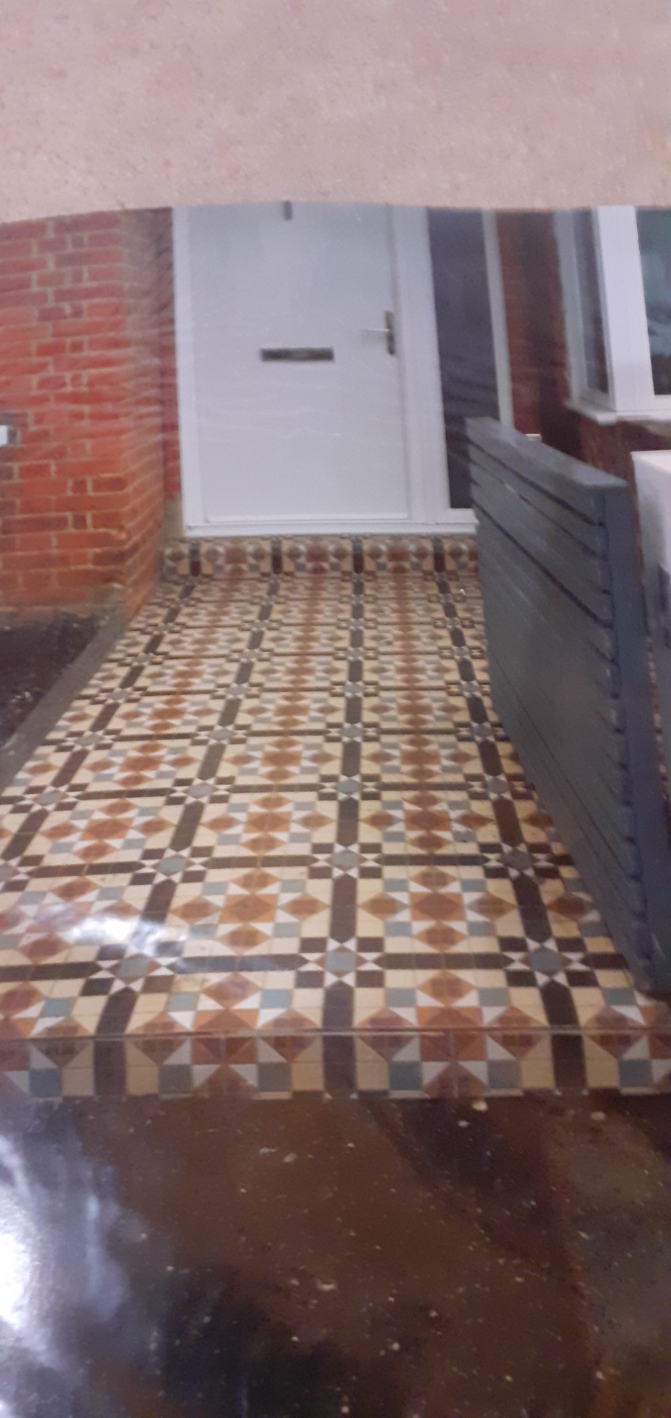 Ułożona, wzorzysta posadzka z płytek ceramicznych przed wejściem do domu z białą stolarką drzwiową i fragmentem muru z czerwonej cegły.