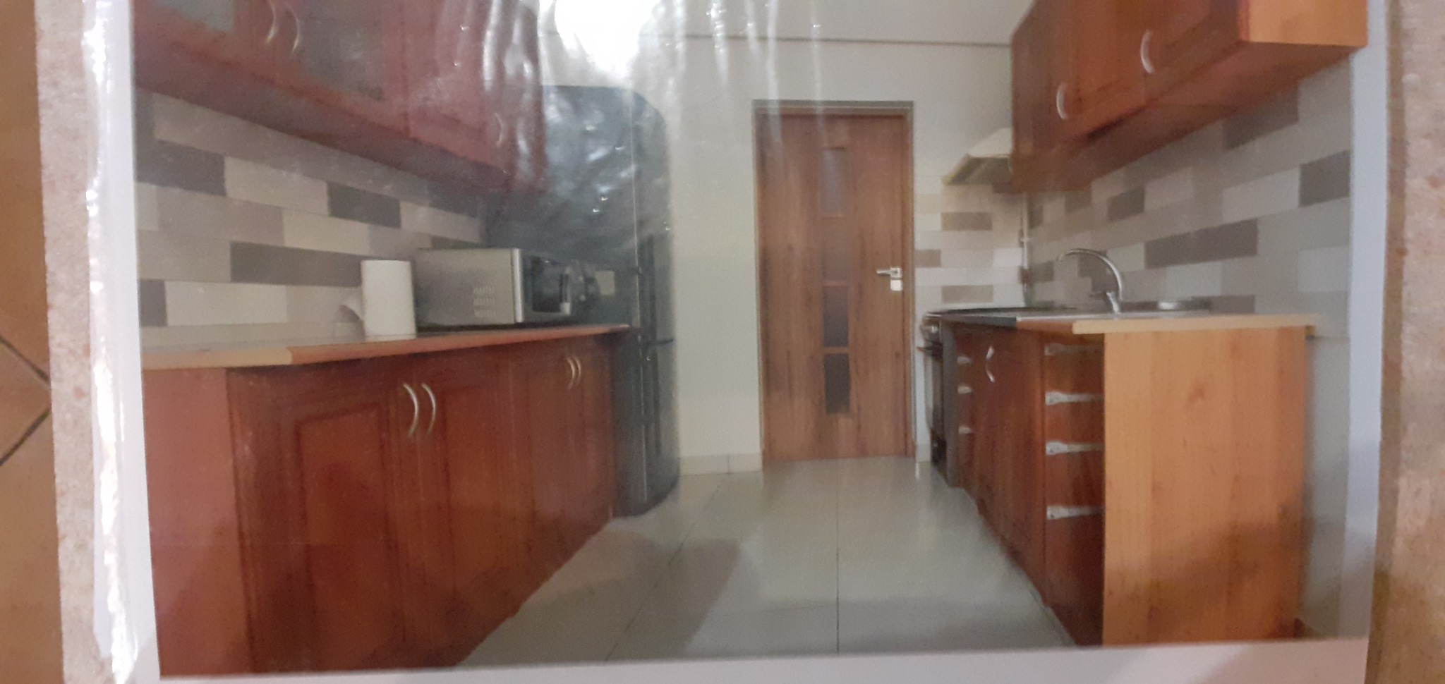 Ustawienie kuchni z drewnianymi szafkami, szarymi płytkami na ścianie, z widocznym zlewem i kuchenką, sfotografowane przez folię ochronną.