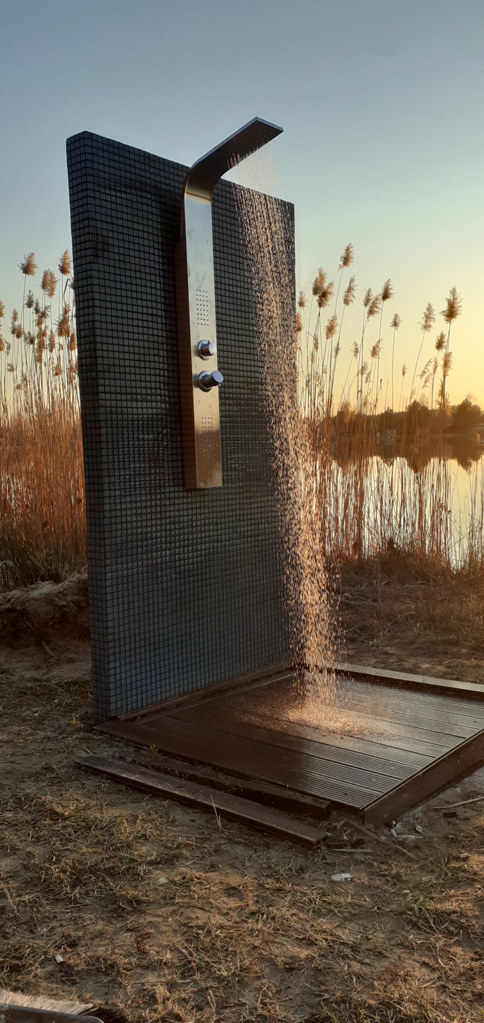 Zewnętrzny prysznic z deszczownicą, obłożony mozaiką, na tle jeziora i trzcin w promieniach zachodzącego słońca.