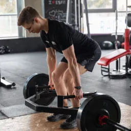 Młody mężczyzna w okularach wykonuje martwy ciąg z użyciem sztangi typu trap bar w siłowni, widoczny napis 'I LOVE ESFIT' na tablicy.