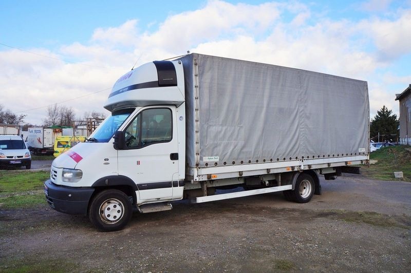 Biały samochód dostawczy z plandeką na szarym podłożu, widok z boku, lekko pochmurne niebo w tle.