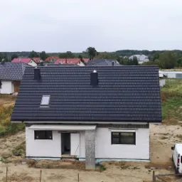 Nowy, grafitowy dach z dachówki ceramicznej na surowym budynku jednorodzinnym z widocznym oknem dachowym i kominem, na tle innych domów i zieleni, z samochodem dostawczym w pobliżu.