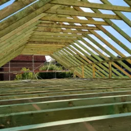 Konstrukcja drewniana w trakcie budowy, widok od wewnątrz, elementy więźby dachowej zaimpregnowane na zielono, w tle budynek z czerwonej cegły i rusztowanie.