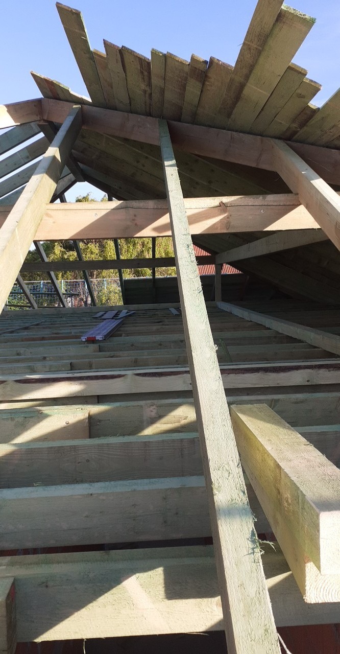 Konstrukcja drewniana dachu w trakcie budowy, widoczne elementy więźby dachowej i deski szczytowe z nieregularnymi krawędziami, jasne drewno w surowym stanie, w tle zieleń drzew.