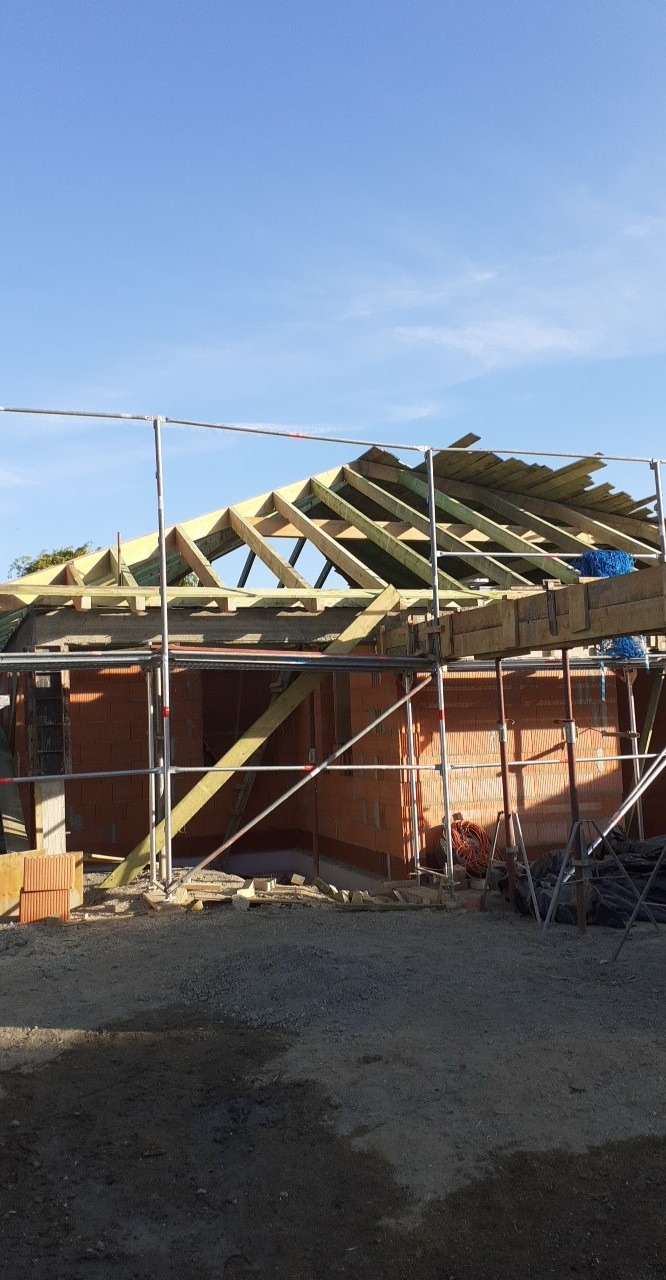 Wznoszenie więźby dachowej na murowanym budynku jednorodzinnym, widoczne rusztowanie, niebieska siatka zabezpieczająca i częściowo ułożone deski poszycia dachu.