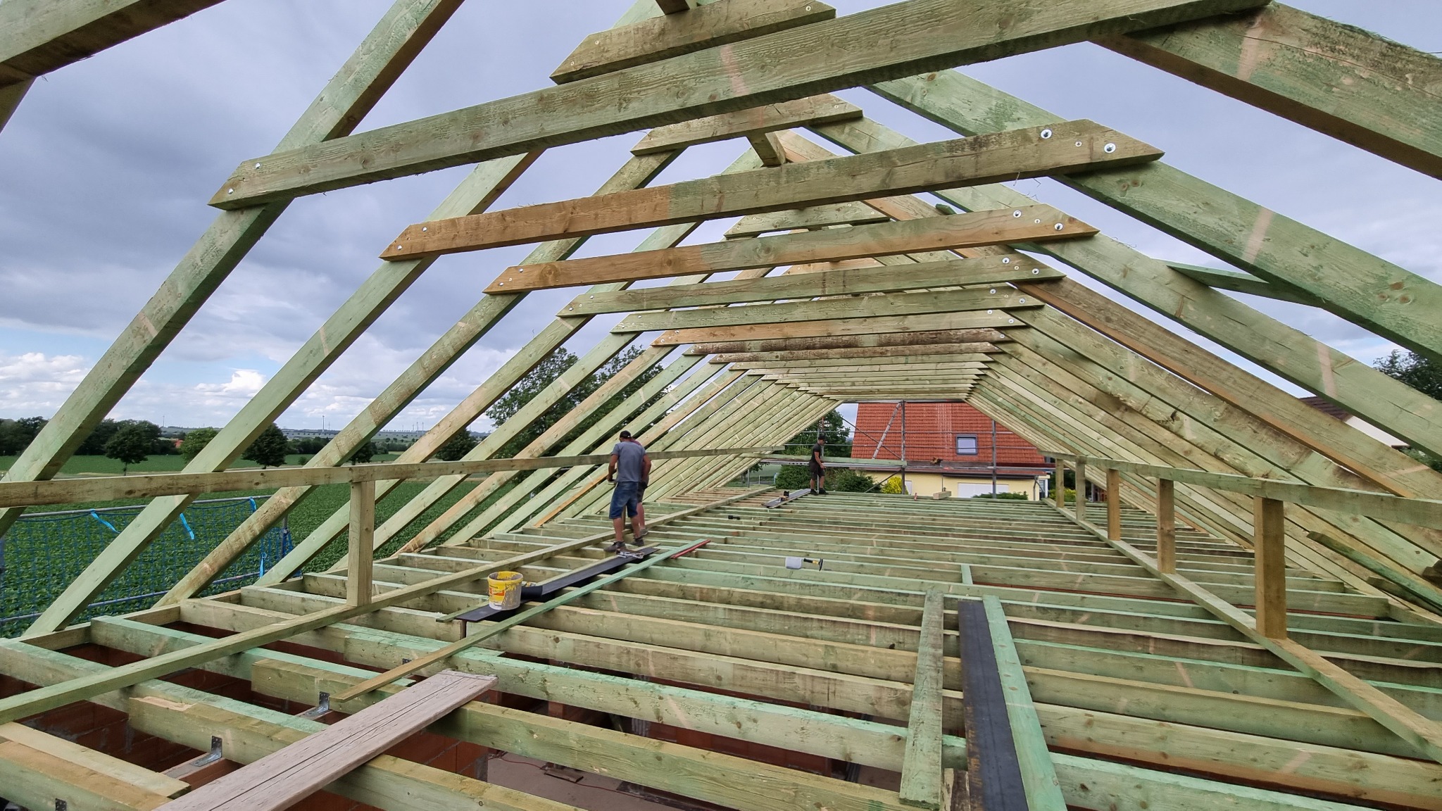 Konstrukcja drewniana dachu w trakcie montażu, widok z góry na robotników pracujących na ażurowej więźbie, z widocznymi elementami mocującymi i fragmentem krajobrazu w tle.