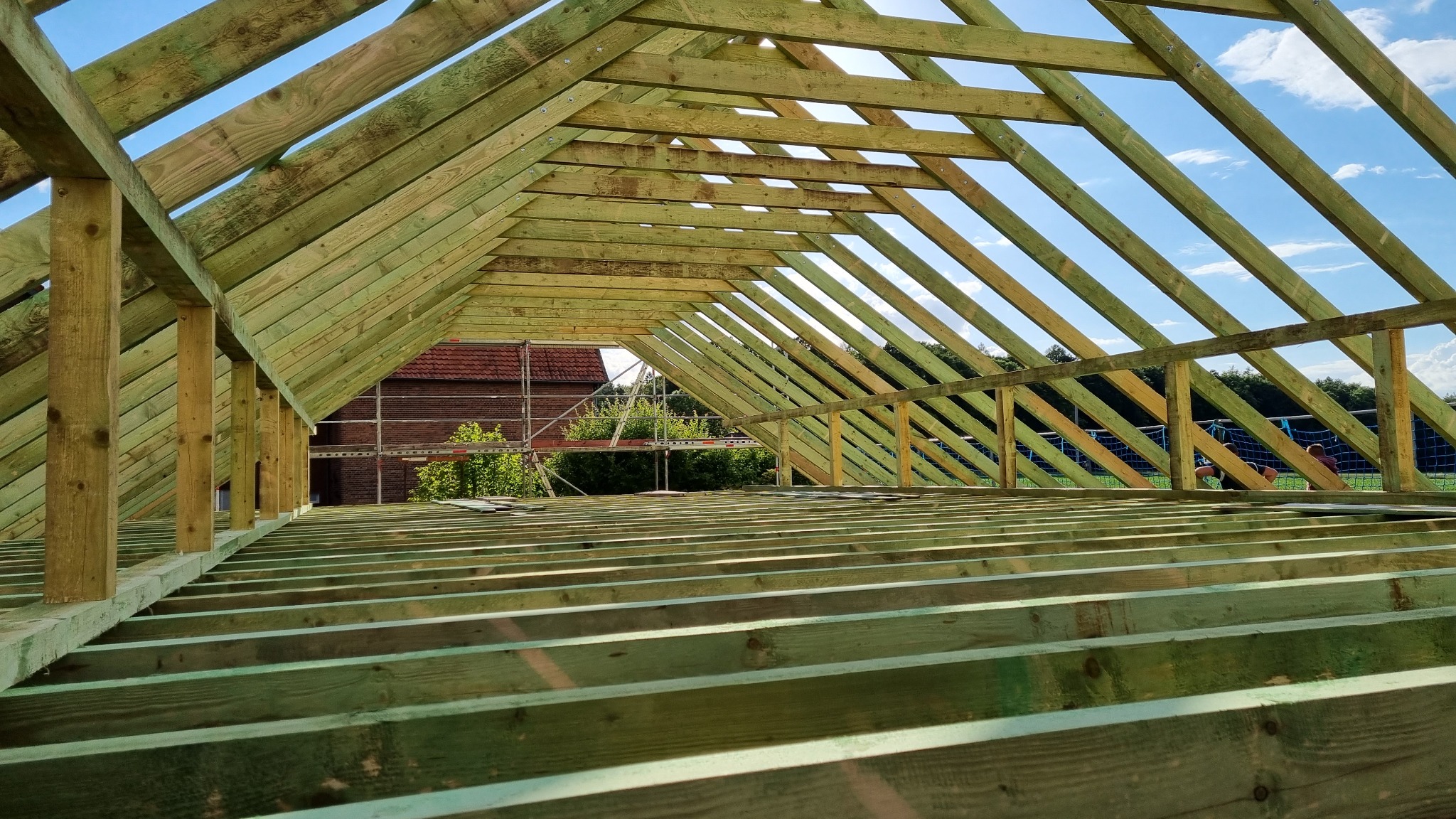 Konstrukcja drewniana w trakcie budowy, widok od wewnątrz, elementy więźby dachowej zaimpregnowane na zielono, w tle budynek z czerwonej cegły i rusztowanie.
