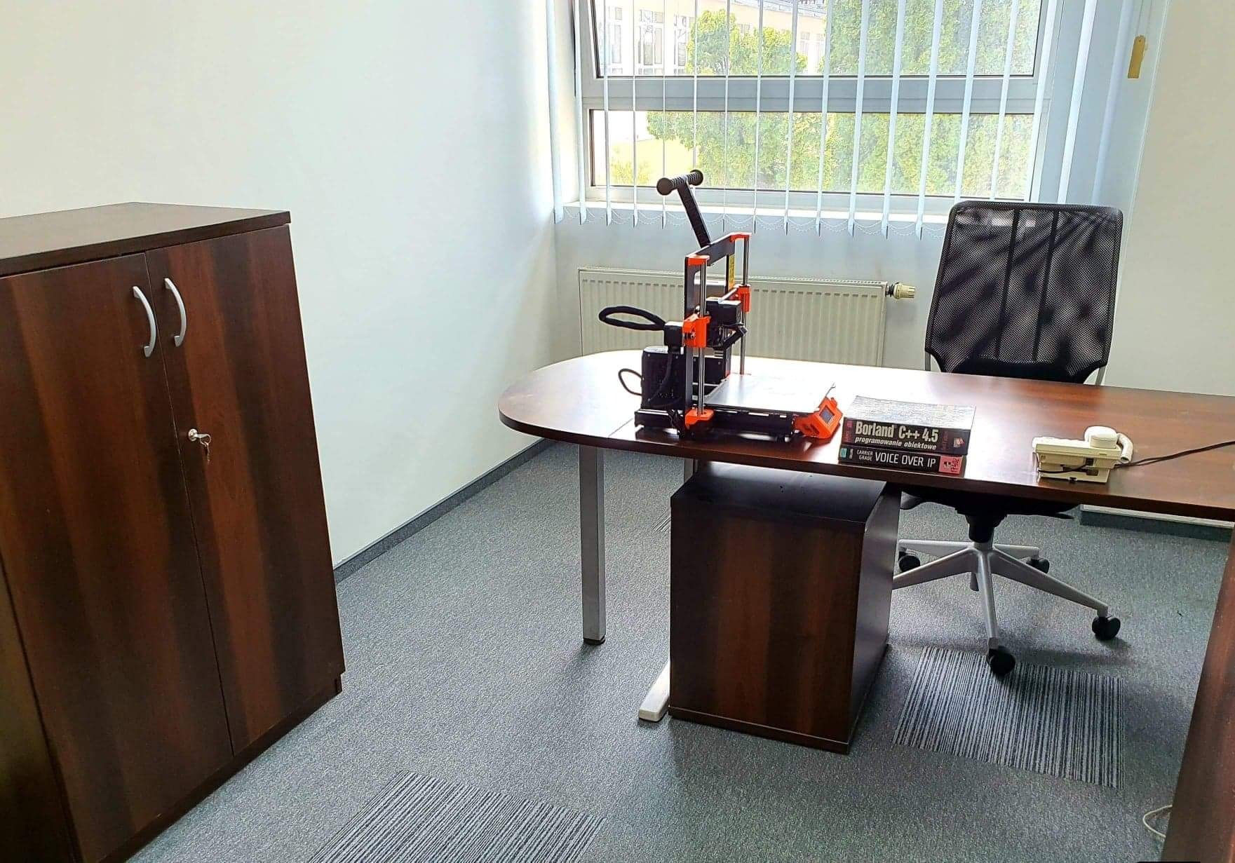Biuro z szarą wykładziną dywanową w jodełkę, biurkiem z drukarką 3D, szafką i krzesłem biurowym na kółkach.