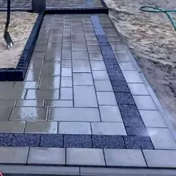 Świeżo ułożona, wilgotna kostka brukowa z ciemnoszarym paskiem dekoracyjnym z drobnego kamienia, widoczna łopata i fragment węża ogrodowego.