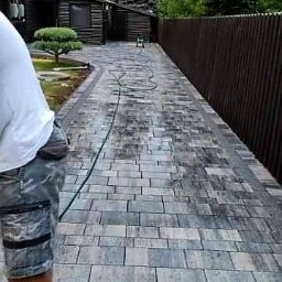 Fabian Patalon - Świeżo ułożona, szara kostka brukowa na podjeździe, delikatnie zwilżana wodą z węża ogrodowego, widoczny fragment osoby w szortach i białej koszulce, brązowe ogrodzenie w tle.