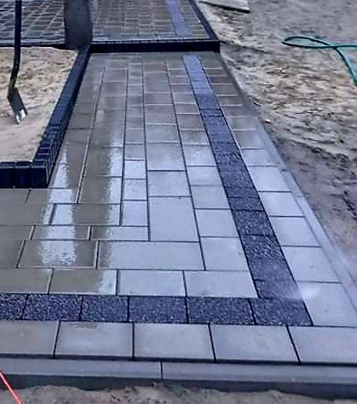 Świeżo ułożona, wilgotna kostka brukowa z ciemnoszarym paskiem dekoracyjnym z drobnego kamienia, widoczna łopata i fragment węża ogrodowego.