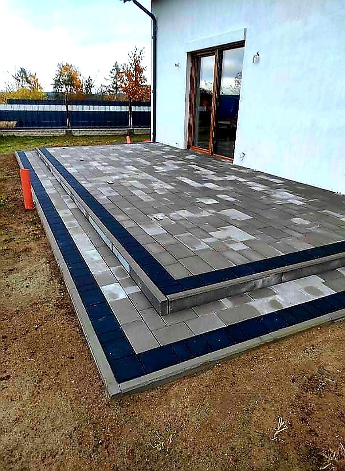 Nowoczesny taras z kostki brukowej w odcieniach szarości z ciemnoniebieskim obramowaniem schodów, przylegający do białego budynku z brązowymi drzwiami tarasowymi, widok z dołu.