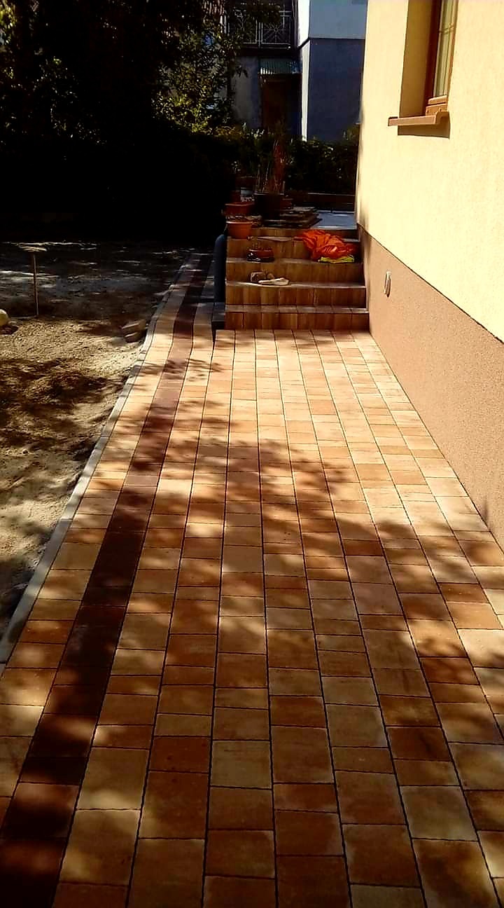 Nowo ułożona ścieżka z kostki brukowej w odcieniach brązu prowadząca do schodów wejściowych przy budynku o beżowej elewacji, częściowo zacieniona przez drzewa.