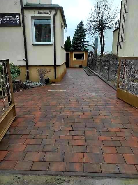 Ułożona, czerwono-brązowa kostka brukowa na podjeździe, widok zza otwartej bramy, budynki po bokach, tabliczka 'Fryzjer Męski' na ścianie.