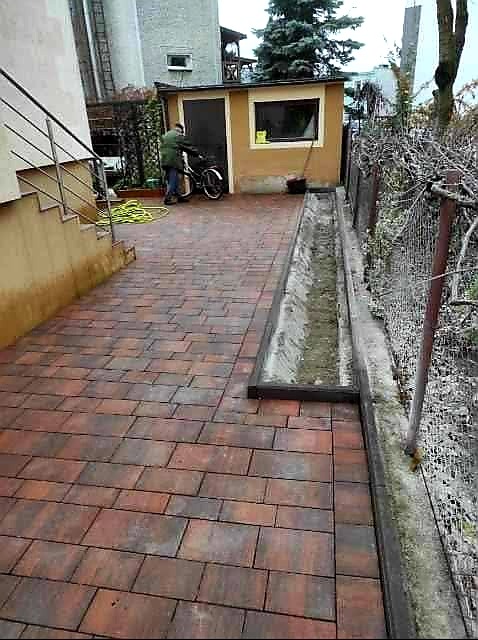 Ułożona kostka brukowa w odcieniach czerwieni i brązu prowadząca do garażu, widoczny odpływ liniowy, rower i osoba w zielonej kurtce.