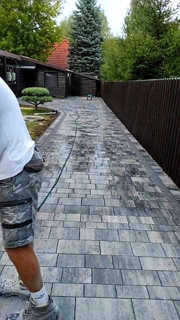 Świeżo ułożona, szara kostka brukowa na podjeździe, delikatnie zwilżana wodą z węża ogrodowego, widoczny fragment osoby w szortach i białej koszulce, brązowe ogrodzenie w tle.