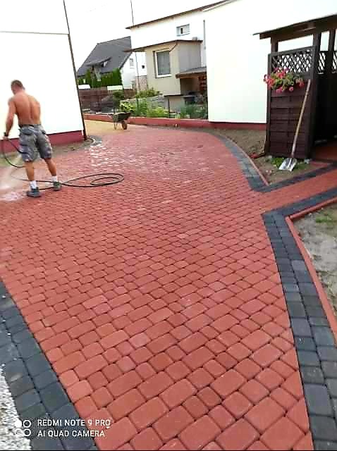 Czerwona kostka brukowa z czarnym obramowaniem, czyszczona myjką ciśnieniową przez mężczyznę bez koszulki na tle domu jednorodzinnego.