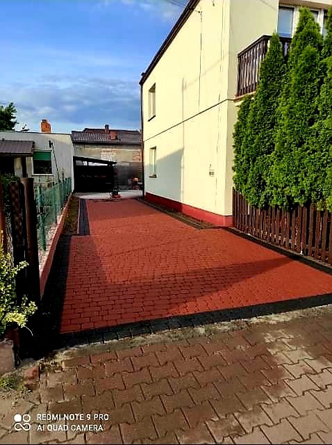 Czerwona kostka brukowa ułożona na podjeździe do domu, obrzeża wykończone czarnym brukiem, w tle budynek z balkonem i zielone tuje.