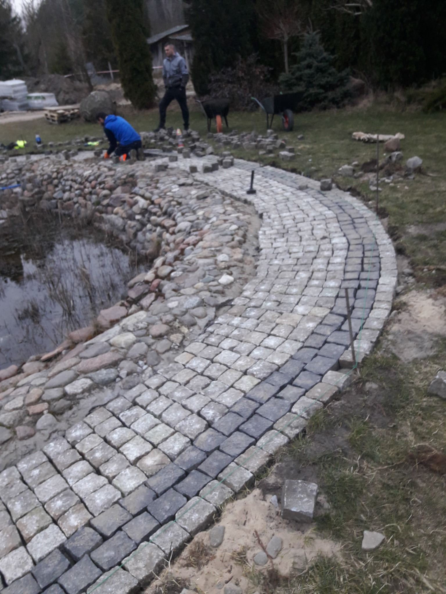 Ścieżka z kostki granitowej układana wokół oczka wodnego, widoczni dwaj pracownicy, jeden w trakcie pracy, drugi nadzorujący, w tle taczki i palety z kostką.