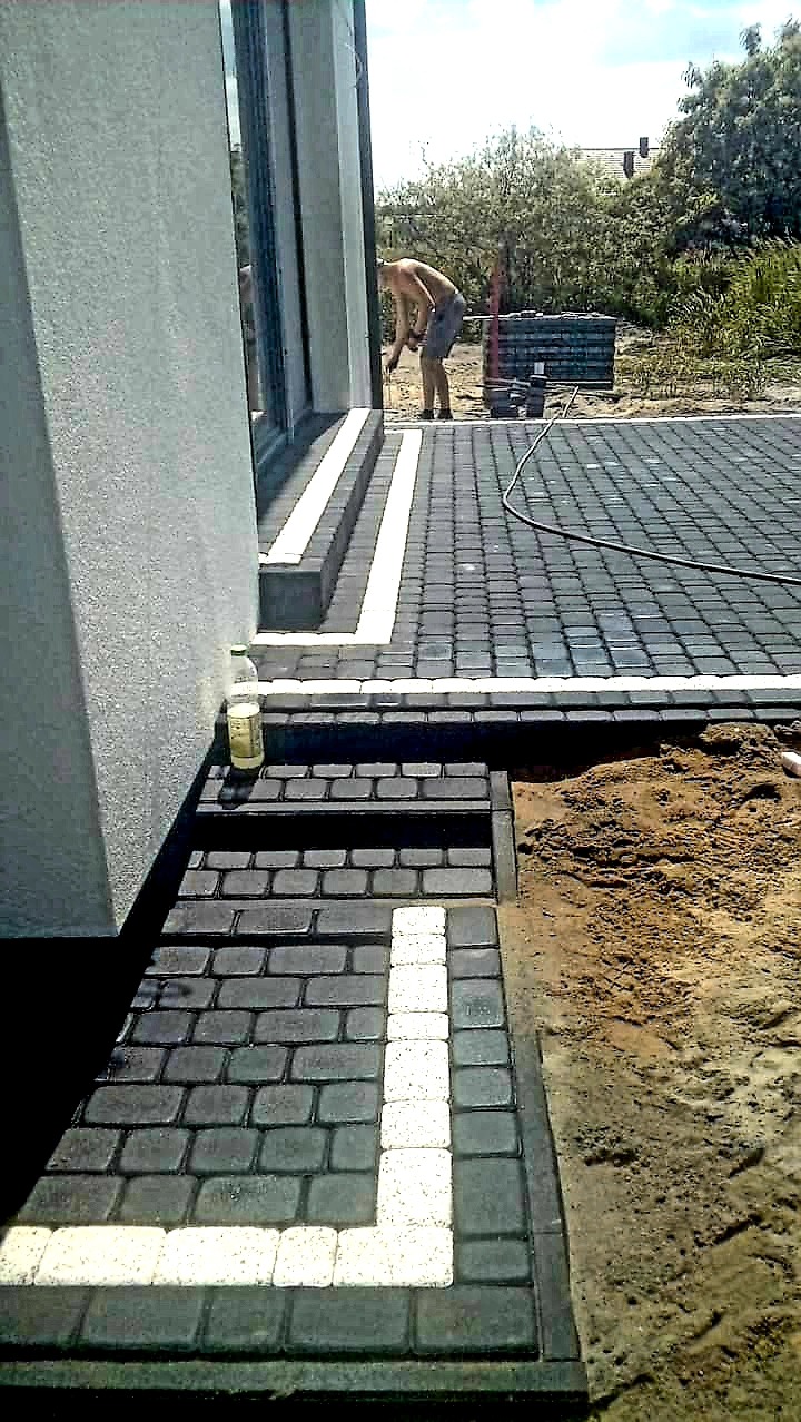 Etap układania ciemnoszarej kostki brukowej z białymi akcentami na schodach i podjeździe przed budynkiem, widoczny pracownik w tle.