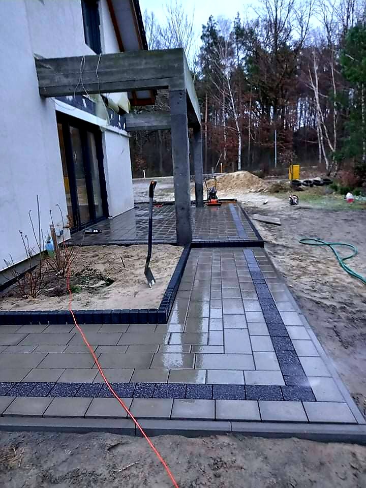 Świeżo ułożona, mokra kostka brukowa w odcieniach szarości z ciemnymi obrzeżami, prowadząca do domu z betonowym zadaszeniem tarasu; widoczna łopata i zagęszczarka w tle.