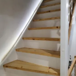 Bieg schodów z drewnianymi stopniami i białymi podstopnicami oświetlony listwą LED, ażurowa balustrada po prawej stronie.