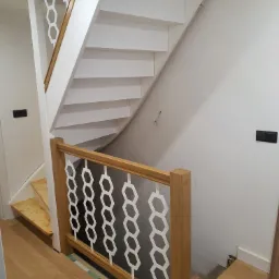 Białe, zabiegowe schody z drewnianą poręczą i nowoczesną balustradą z białymi, heksagonalnymi wycięciami, widoczne niedokończone wykończenie podłogi.
