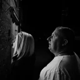 Czarno-białe zdjęcie przedstawia kobietę z blond włosami i mężczyznę z krótkimi włosami, oboje patrzą w górę na budynek z cegły w słabym oświetleniu.