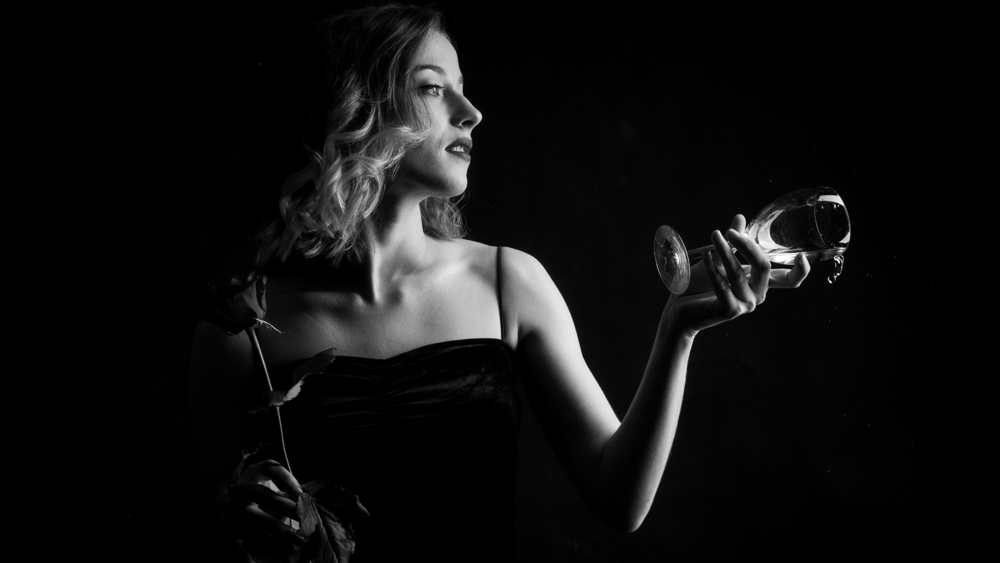 Portret kobiety w czarno-białej tonacji, trzymającej różę w jednej ręce i przechylony kieliszek z kroplami w drugiej, na ciemnym tle.