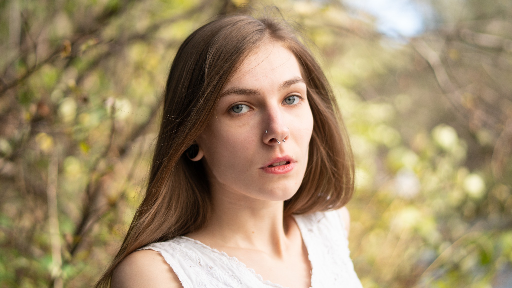 Portret młodej kobiety z piercingiem nosa i tunelem w uchu, w białej bluzce na tle rozmytej zieleni.