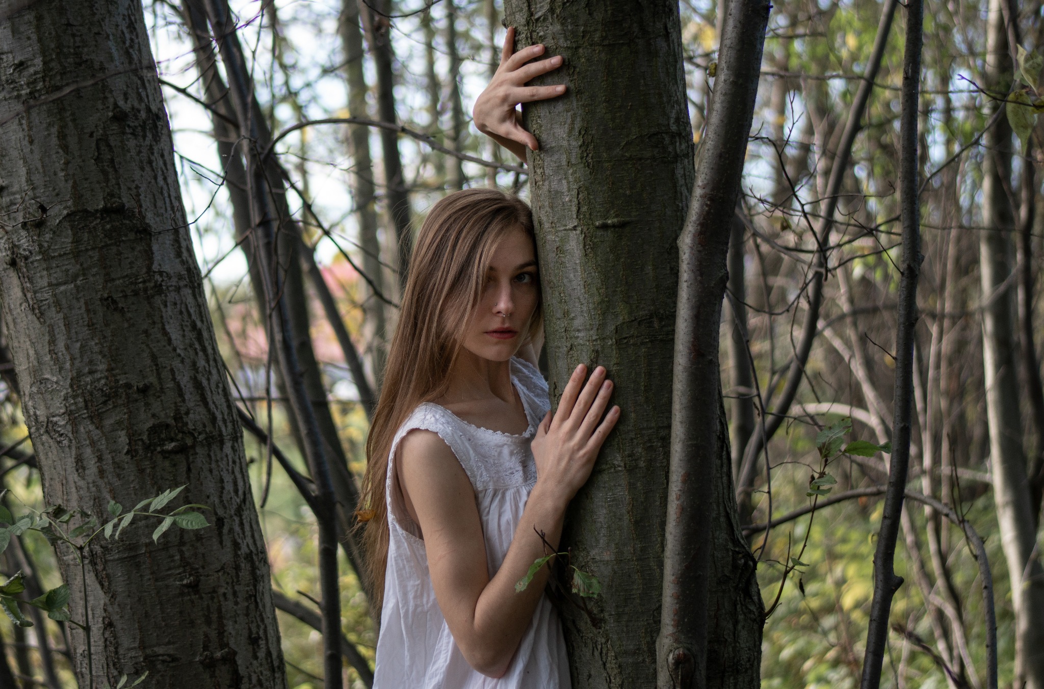 Portret kobiety w białej sukience, opierającej się o drzewo w lesie. Naturalne światło, leśna sceneria, melancholijny wyraz twarzy, delikatny makijaż, artystyczne ujęcie.