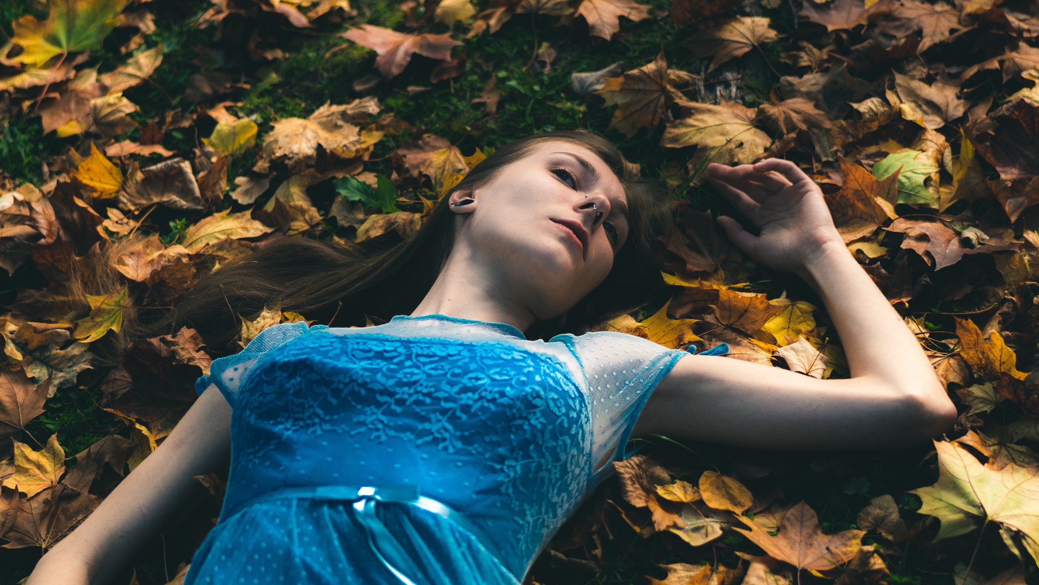Młoda kobieta w błękitnej sukience leży na ziemi pokrytej jesiennymi liśćmi, z głową odchyloną do tyłu i ręką wyciągniętą w górę, skierowana twarzą do góry.