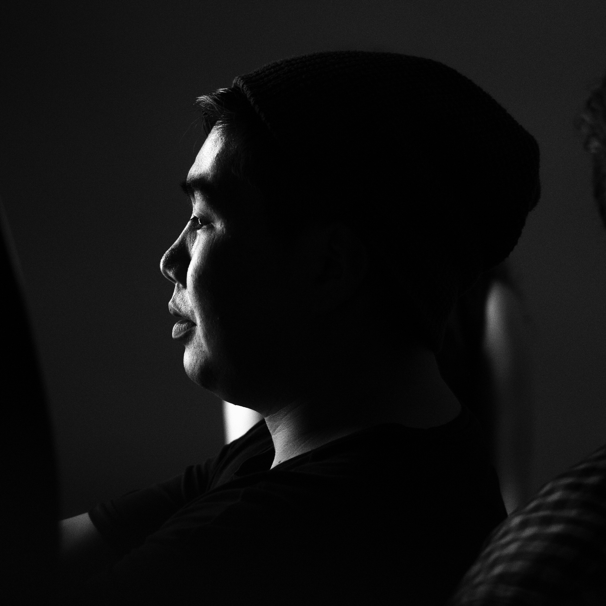 Czarno-białe, profilowe ujęcie twarzy mężczyzny w czapce, oświetlone bocznym, kontrastowym światłem, z widocznymi detalami rysów twarzy.