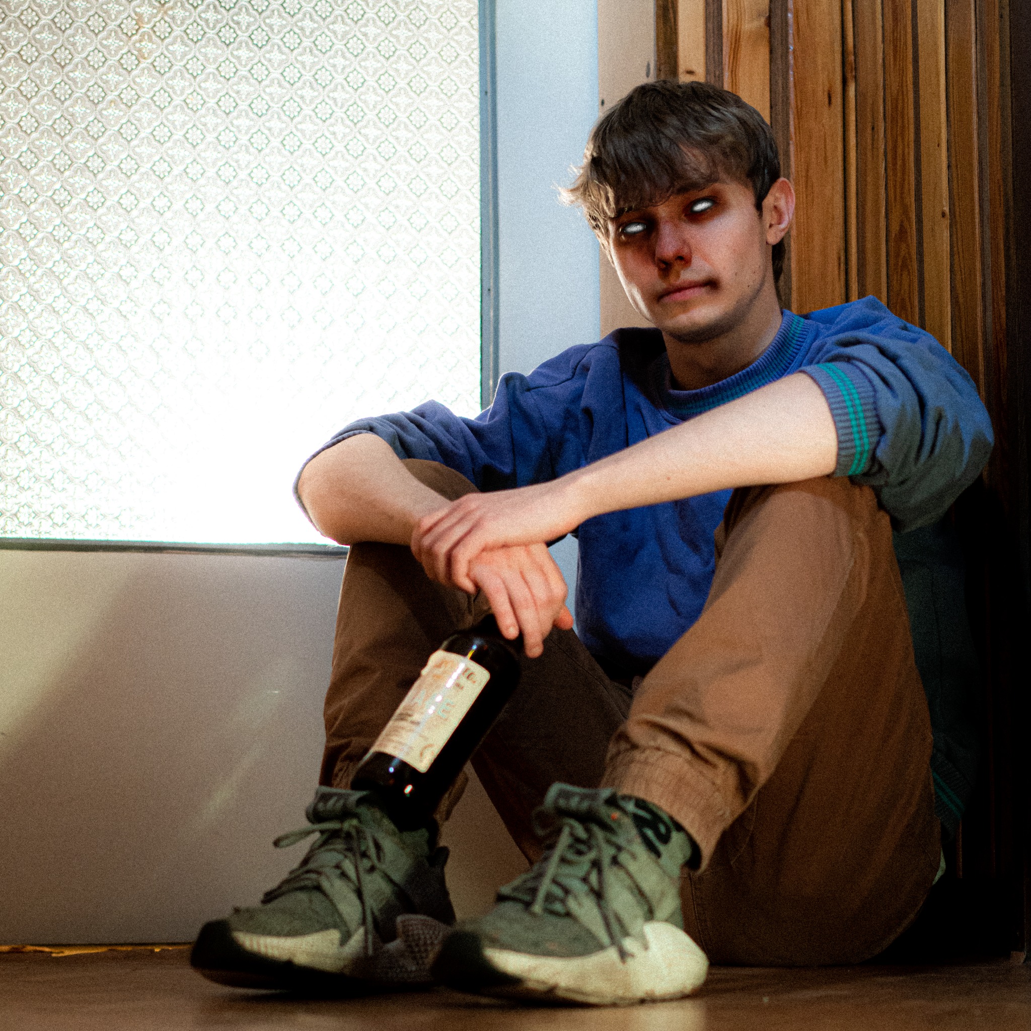 Młody mężczyzna siedzi na podłodze przy oknie, trzymając butelkę wina, z niepokojącym spojrzeniem i białymi soczewkami w oczach.