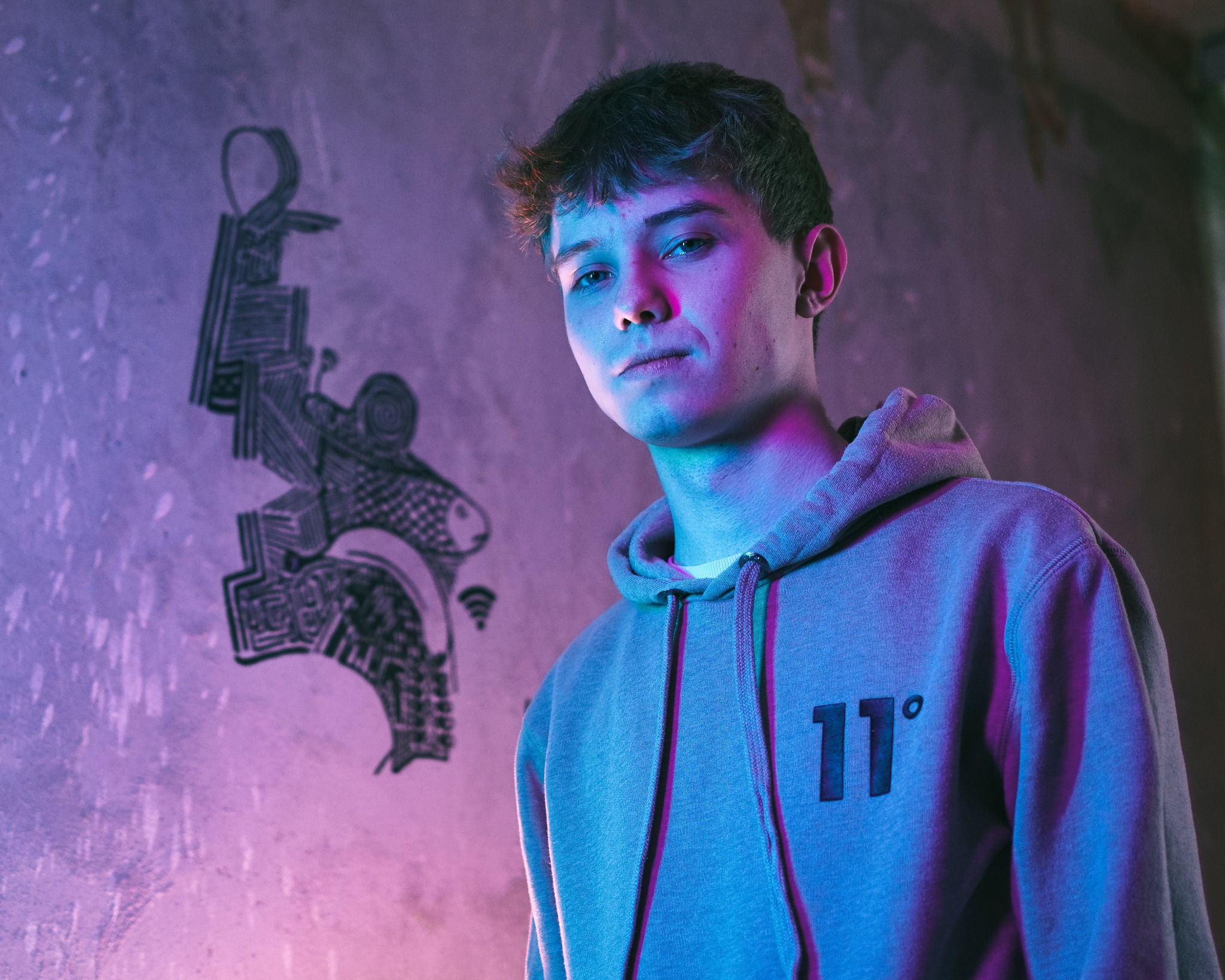 Portret młodego mężczyzny w szarej bluzie z kapturem na tle betonowej ściany z abstrakcyjnym graffiti, oświetlony neonowym, niebiesko-fioletowym światłem.