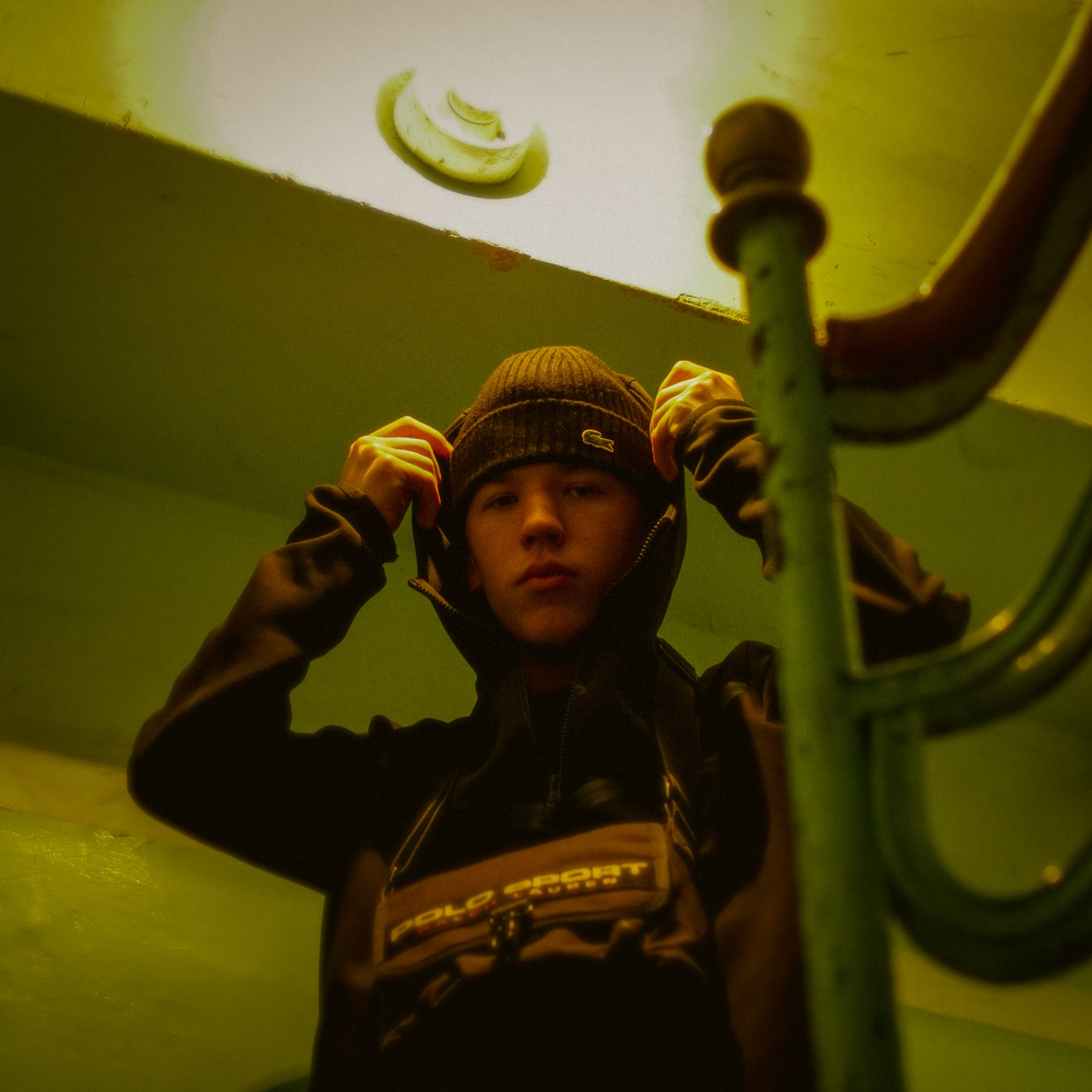 Młody mężczyzna w czapce i bluzie z kapturem, pozujący na tle schodów z metalową barierką i sufitowym oświetleniem w odcieniach sepii.