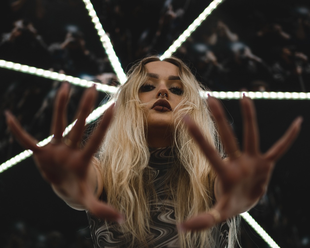 Portret kobiety z blond włosami, piercingiem w nosie i pierścionkami na palcach, wyciągającej dłonie w stronę obiektywu na tle geometrycznej instalacji świetlnej.