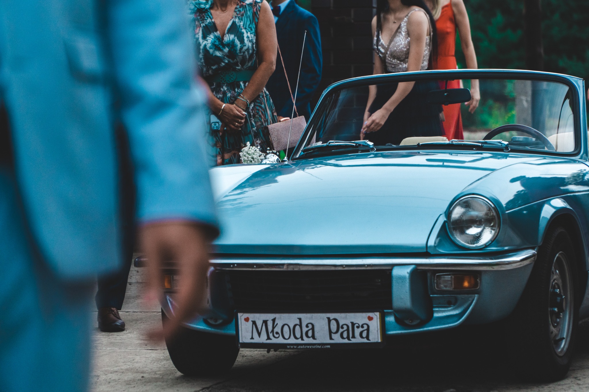 Błękitny kabriolet z tablicą 'Młoda Para' na tle gości weselnych, ujęcie z bliska, z widocznym fragmentem osoby w niebieskim garniturze na pierwszym planie.