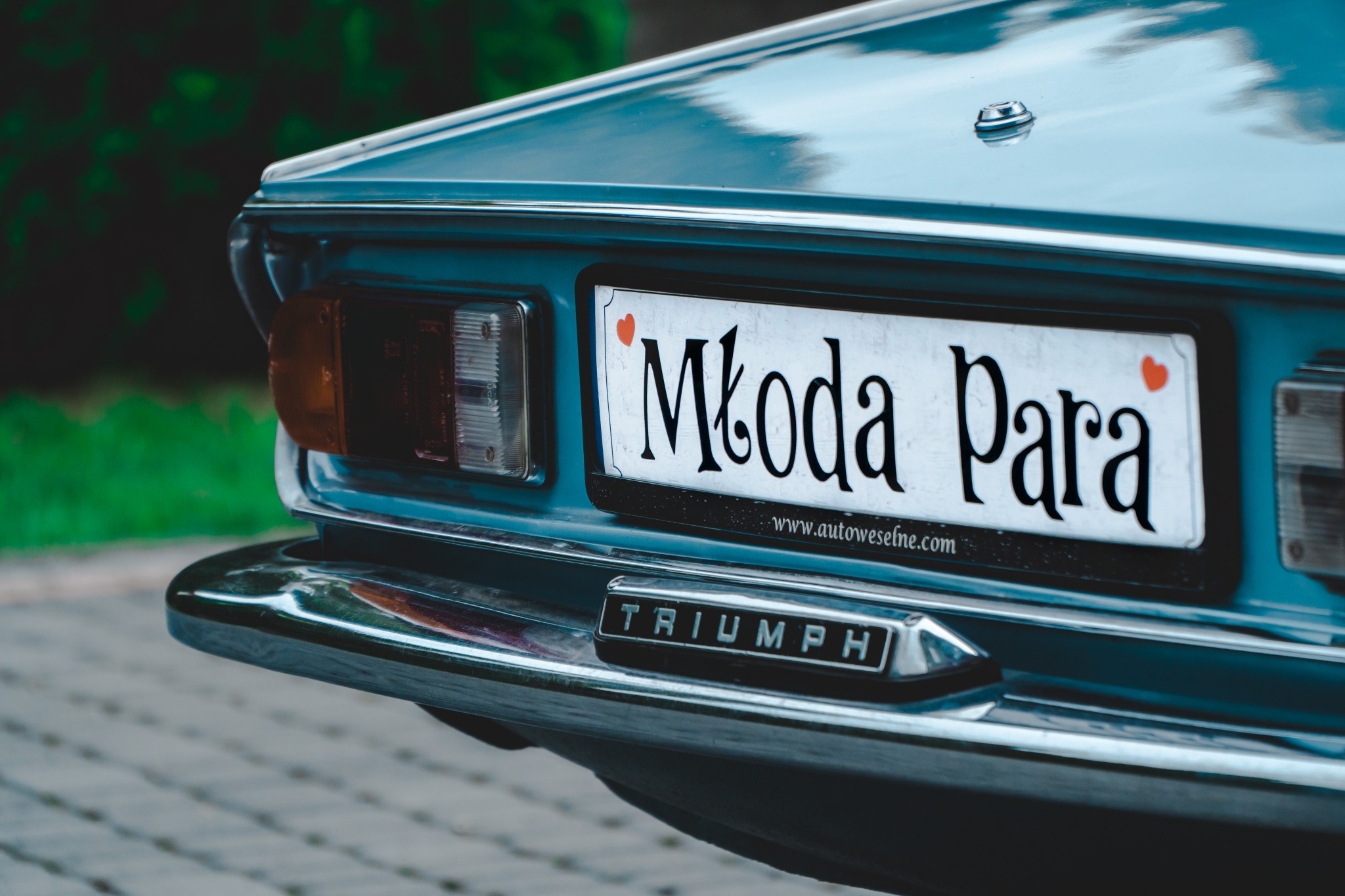 Fragment niebieskiego samochodu marki Triumph z tablicą rejestracyjną 'Młoda Para' ozdobioną małymi czerwonymi serduszkami, widoczne tylne światło i chromowany zderzak.