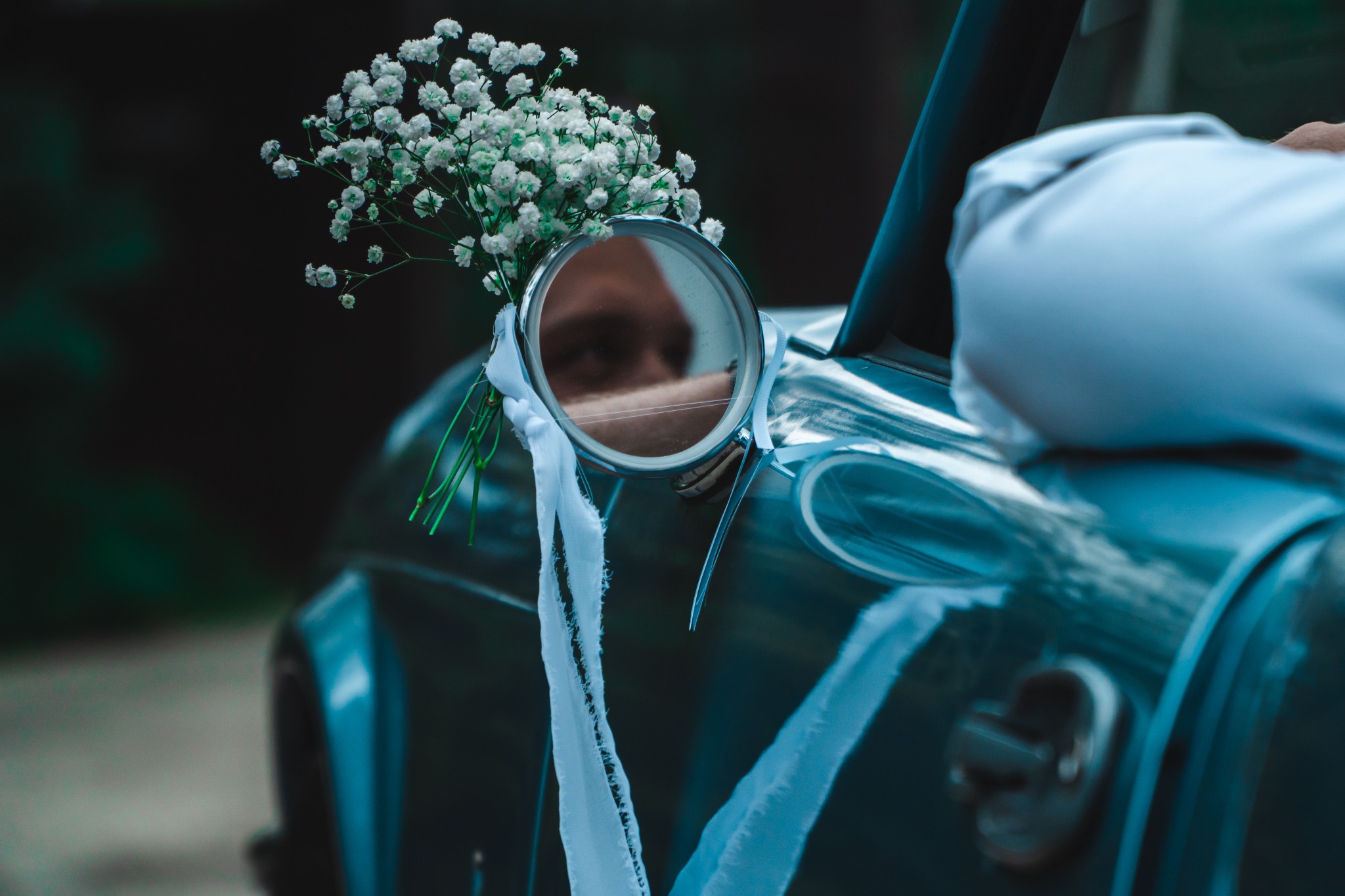 Udekorowany lusterkiem wstecznym samochód ślubny z bukiecikiem gipsówki i białymi wstążkami, odbicie twarzy w lustrze.