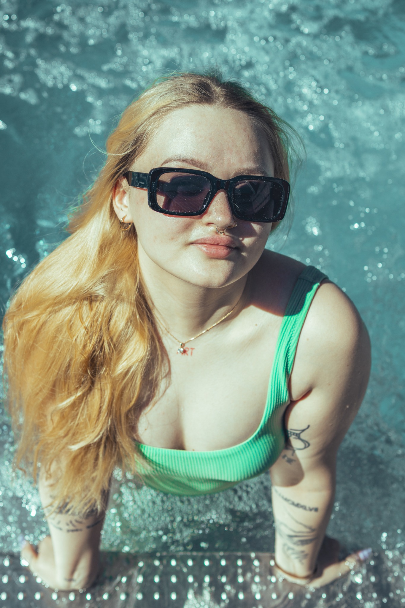 Portret kobiety w okularach przeciwsłonecznych, w zielonym stroju kąpielowym, zanurzonej w wodzie, z widocznymi tatuażami na ramionach.